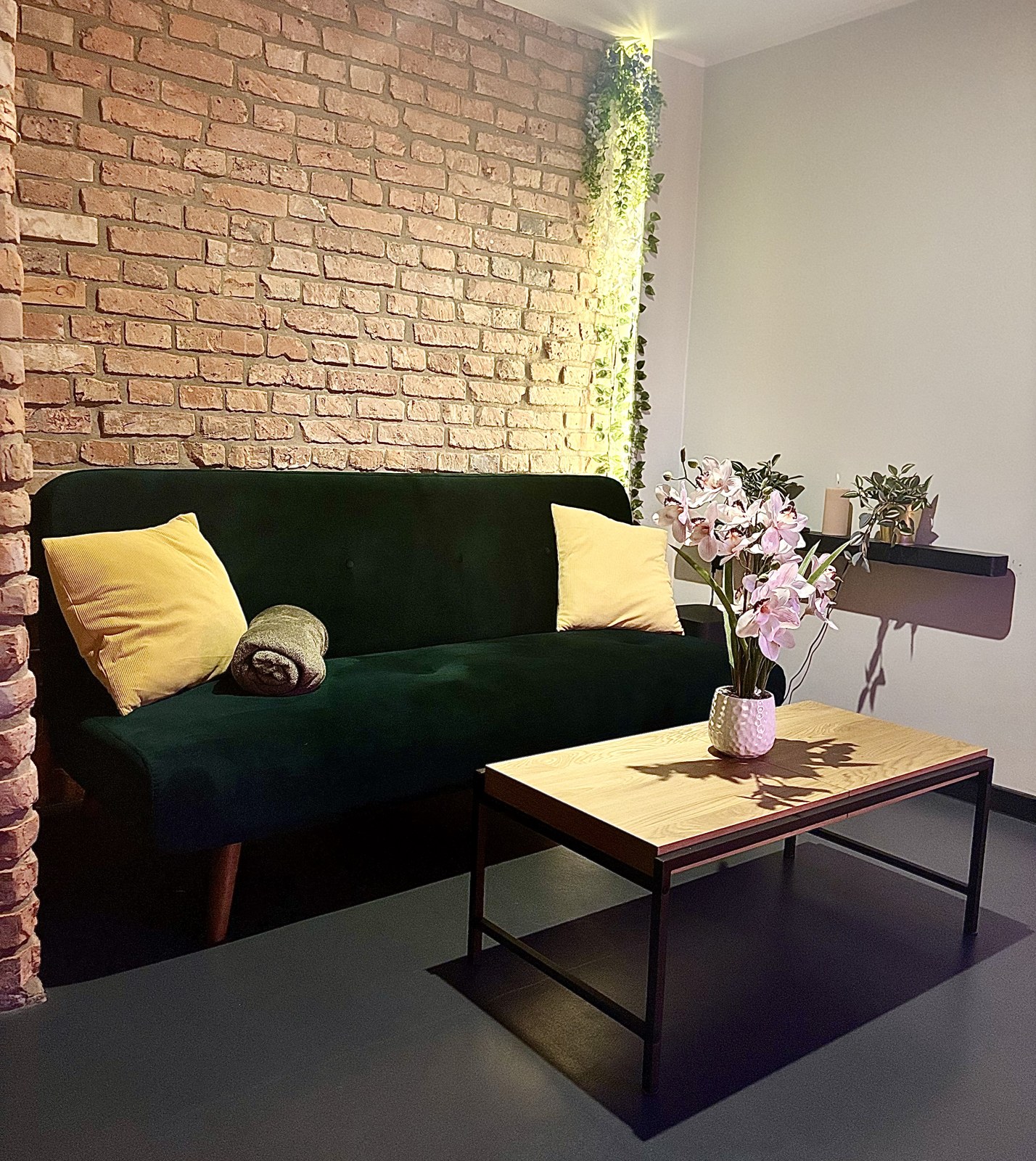 Living room corner with green velvet sofa, yellow cushions and an orchid against exposed brick — Property Presentation Reset, Poznań apartment